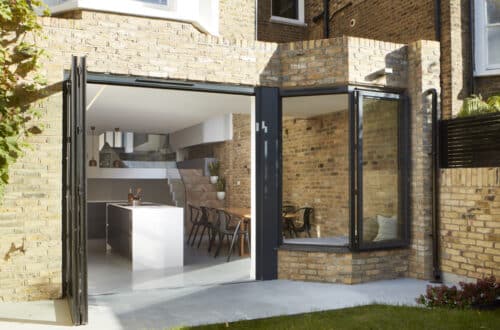 Brick exterior with folding glass doors opening to a modern kitchen and dining area with a large island and wooden table.