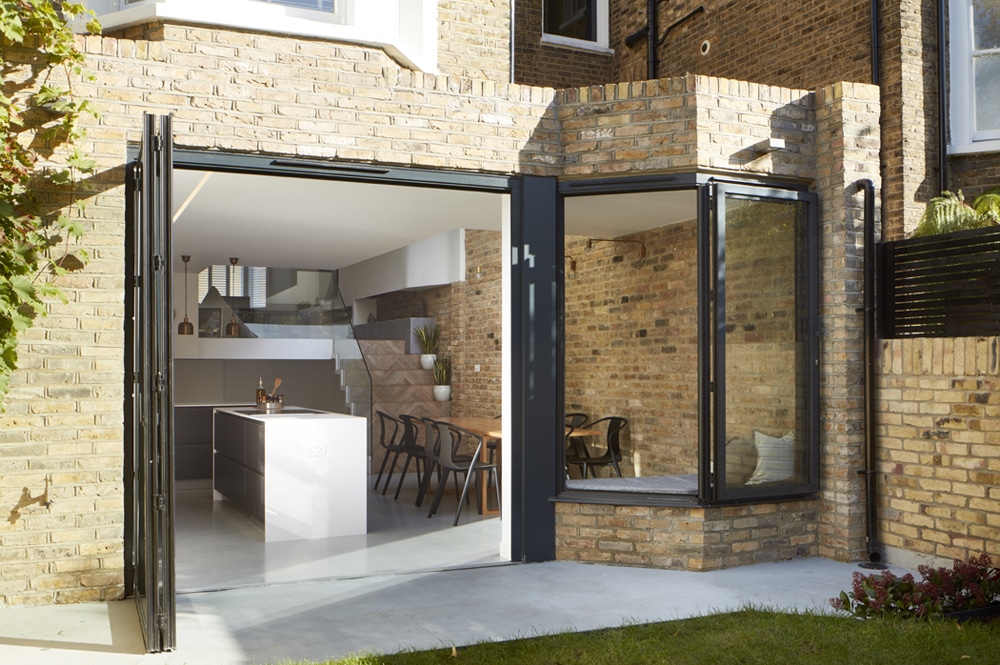 Brick exterior with folding glass doors opening to a modern kitchen and dining area with a large island and wooden table.