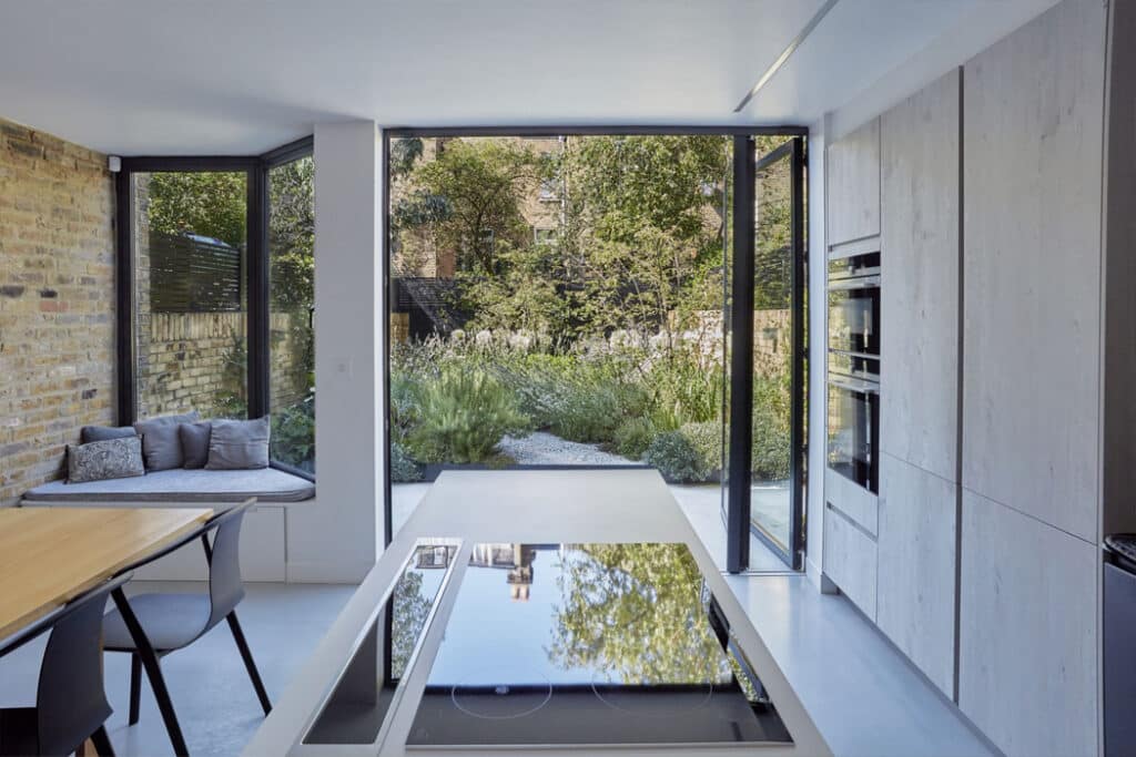 Modern kitchen with built-in appliances, island cooktop, and window seating. Large glass doors open to a garden with lush greenery. Light wood and brick accents.