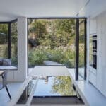 Thumbnail of http://Modern%20kitchen%20with%20built-in%20appliances,%20island%20cooktop,%20and%20window%20seating.%20Large%20glass%20doors%20open%20to%20a%20garden%20with%20lush%20greenery.%20Light%20wood%20and%20brick%20accents.