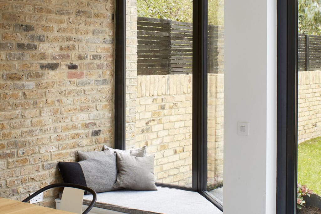 Corner of a room with large glass windows, brick walls, and a small cushioned seating area.