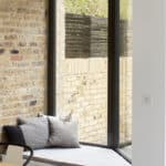 Thumbnail of http://Corner%20of%20a%20room%20with%20large%20glass%20windows,%20brick%20walls,%20and%20a%20small%20cushioned%20seating%20area.