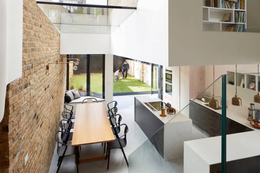 Modern kitchen and dining area with a long table, black chairs, and light wood accents. Glass doors open to a grassy backyard where two people stand. Exposed brick walls and glass railing visible.