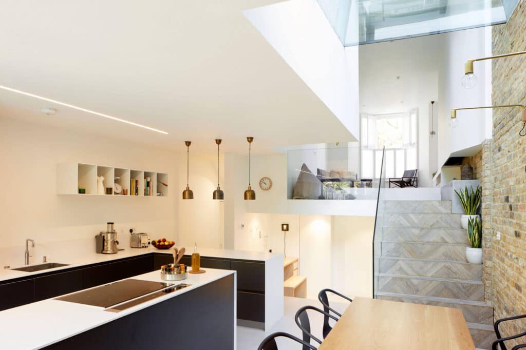 Modern kitchen with black and white cabinetry, island with stovetop, pendant lights, and dining table. Stairs lead to a bright, open living area with a large window and a brick accent wall.