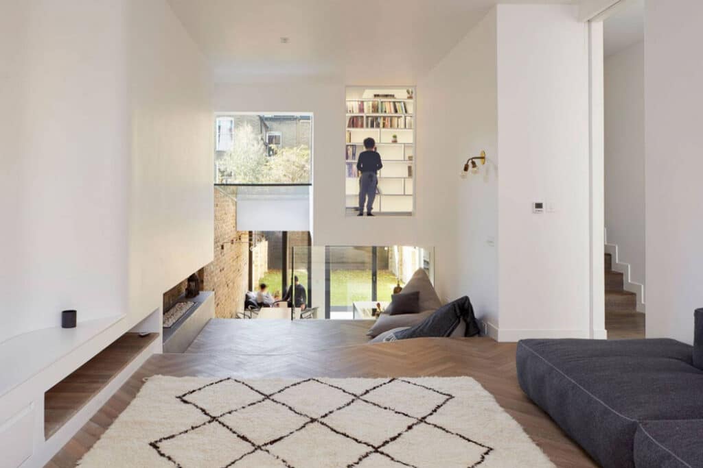 A modern living room with white walls, wooden floor, and a patterned rug. A person stands in a small loft area with a bookshelf, accessed by a ladder. Large windows overlook a garden.