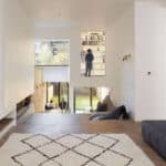 Thumbnail of http://A%20modern%20living%20room%20with%20white%20walls,%20wooden%20floor,%20and%20a%20patterned%20rug.%20A%20person%20stands%20in%20a%20small%20loft%20area%20with%20a%20bookshelf,%20accessed%20by%20a%20ladder.%20Large%20windows%20overlook%20a%20garden.