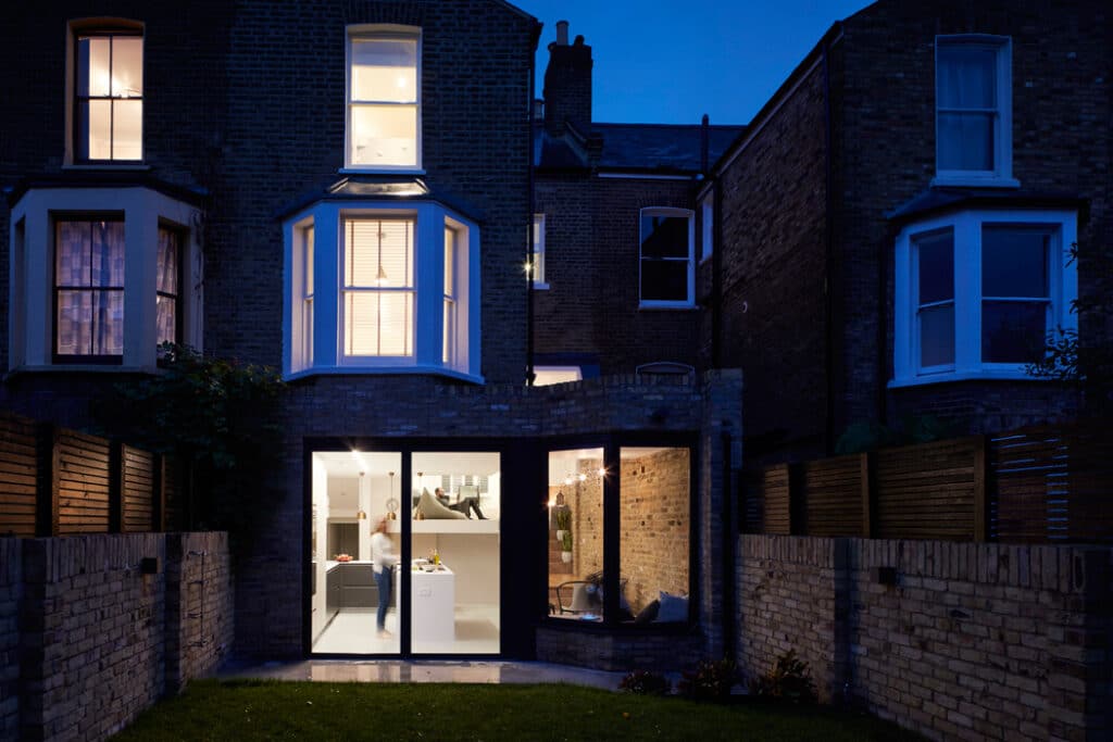 Modern extension with large glass doors on a traditional brick townhouse, interior warmly lit, viewed from the backyard at dusk.
