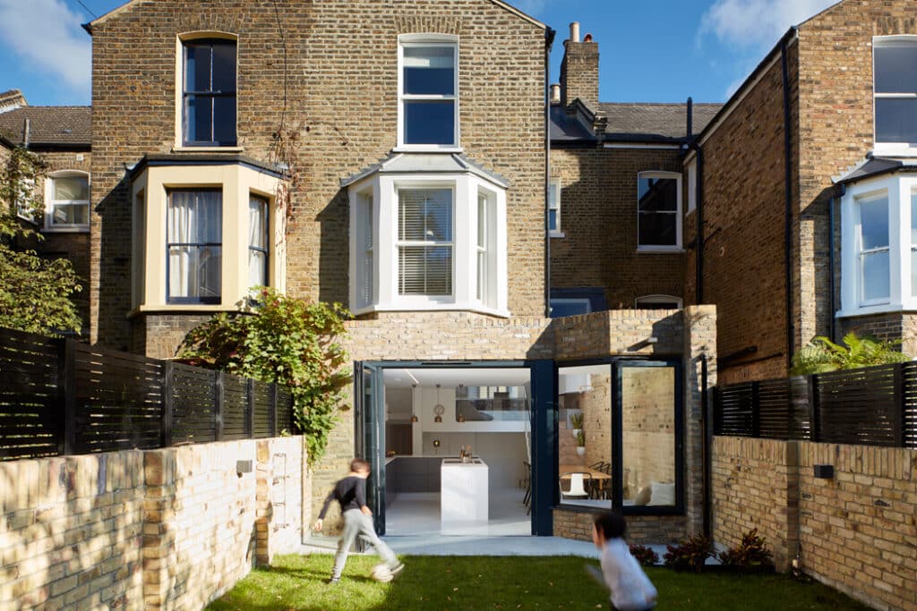 Two children run in the yard of a brick house with large glass doors and windows. The house features a modern extension and a small garden area surrounded by a brick and fence boundary.