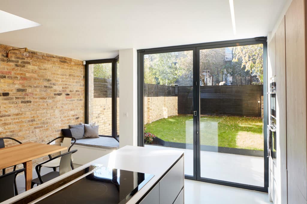 Modern kitchen with an island, brick walls, and a dining table. Large glass doors open to a small grassy backyard with a wooden fence.