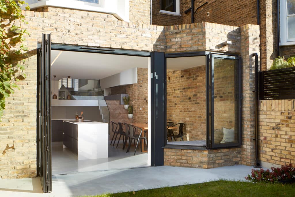 A modern brick house with open bi-fold glass doors leading to a spacious kitchen and dining area. The interior features a white kitchen island and wooden dining table with chairs.