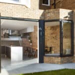 Thumbnail of http://A%20modern%20brick%20house%20with%20open%20bi-fold%20glass%20doors%20leading%20to%20a%20spacious%20kitchen%20and%20dining%20area.%20The%20interior%20features%20a%20white%20kitchen%20island%20and%20wooden%20dining%20table%20with%20chairs.