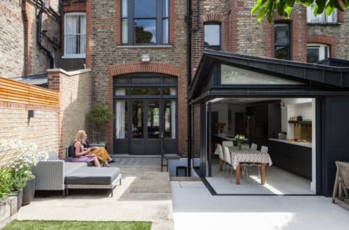A person sits on outdoor furniture in a courtyard beside a brick building. The home features an open glass door leading to a modern kitchen and dining area.