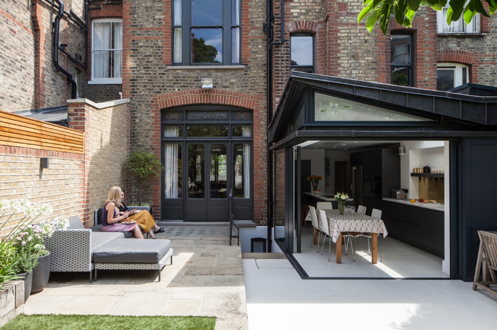 A person sits on outdoor furniture in a courtyard beside a brick building. The home features an open glass door leading to a modern kitchen and dining area.