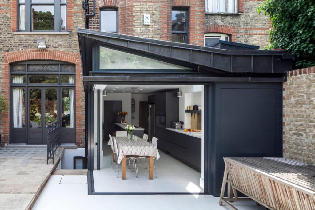 Modern kitchen extension with glass doors, a table set for dining, and sleek cabinetry. The space opens to a courtyard with a wooden bench and brick walls.