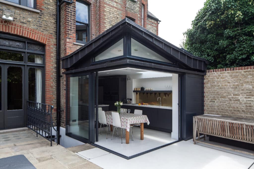 Modern kitchen extension with glass walls attached to a brick house, featuring a dining table with a checkered tablecloth and chairs inside.
