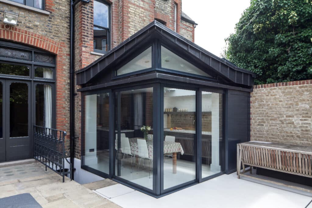 A modern glass conservatory with a dark frame extends from a brick house, featuring a dining table inside and a wooden bench outside.