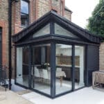 Thumbnail of http://A%20modern%20glass%20conservatory%20with%20a%20dark%20frame%20extends%20from%20a%20brick%20house,%20featuring%20a%20dining%20table%20inside%20and%20a%20wooden%20bench%20outside.