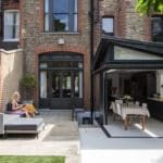 Thumbnail of http://A%20woman%20sits%20on%20outdoor%20furniture%20in%20a%20patio%20area,%20while%20a%20modern%20kitchen%20with%20large%20windows%20is%20visible%20in%20the%20background.