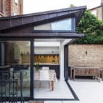 Thumbnail of http://Modern%20house%20extension%20with%20large%20glass%20doors%20and%20triangular%20roof.%20Inside,%20a%20dining%20table%20with%20chairs%20is%20visible.%20Outside,%20a%20wooden%20bench%20sits%20on%20a%20paved%20patio%20surrounded%20by%20brick%20walls%20and%20plants.