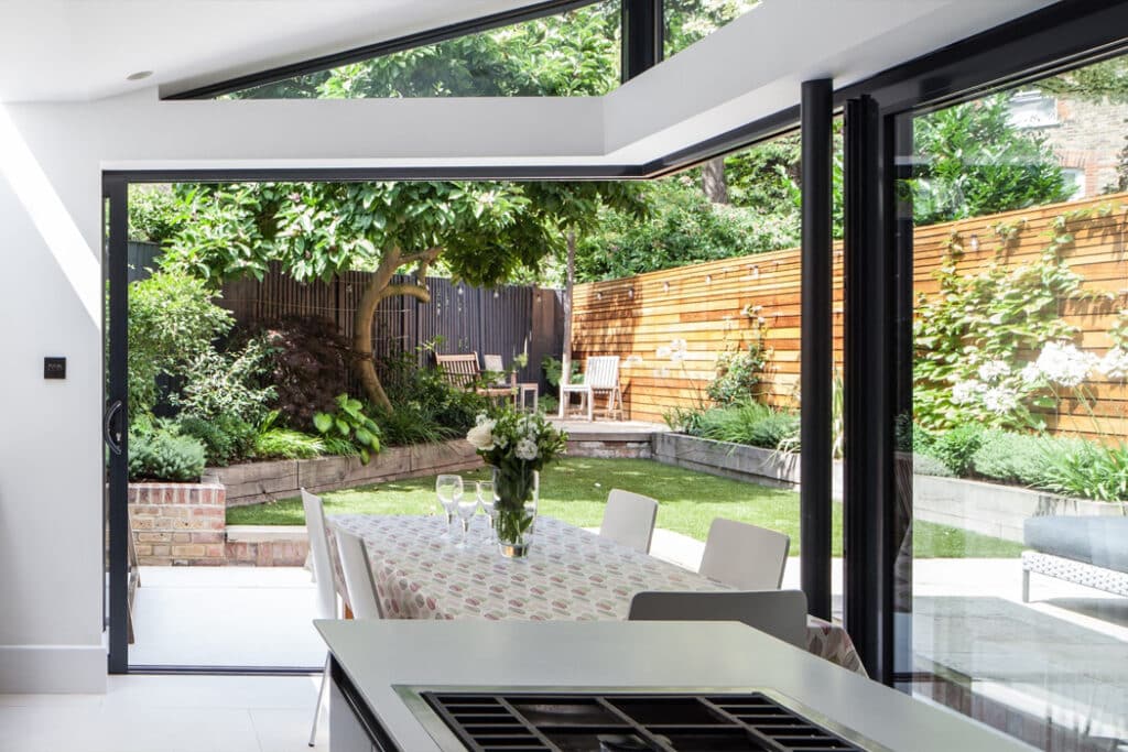 A modern kitchen with a white island overlooks a garden through open glass doors. A table with a patterned cloth and floral centerpiece sits nearby, with chairs outside on the patio.
