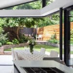 Thumbnail of http://A%20modern%20kitchen%20with%20a%20white%20island%20overlooks%20a%20garden%20through%20open%20glass%20doors.%20A%20table%20with%20a%20patterned%20cloth%20and%20floral%20centerpiece%20sits%20nearby,%20with%20chairs%20outside%20on%20the%20patio.