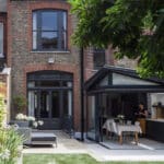 Thumbnail of http://Brick%20house%20with%20a%20modern%20glass%20extension%20featuring%20outdoor%20seating,%20a%20garden%20area%20with%20a%20wooden%20fence,%20and%20trees%20providing%20shade.%20A%20person%20is%20inside%20the%20open%20kitchen.