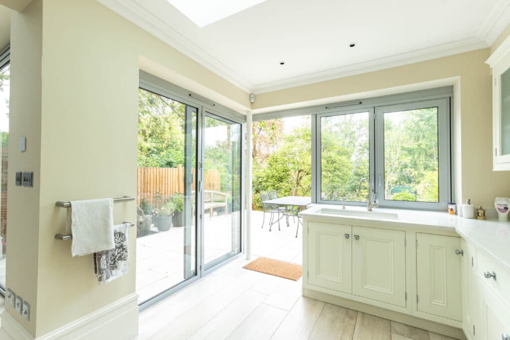 Bright kitchen with light cabinetry, a towel rack, and large sliding glass doors leading to an outdoor patio with a table and chairs. Greenery visible through the glass doors.