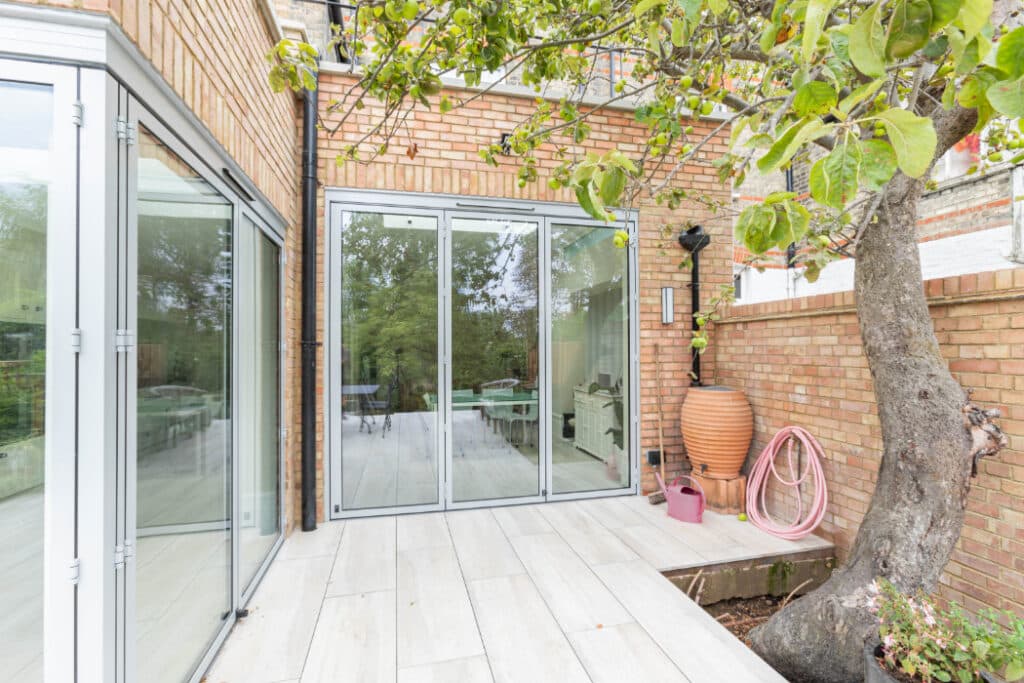 Patio area with sliding glass doors, tree near wall, pink hose, large terracotta pot, and outdoor furniture visible inside.