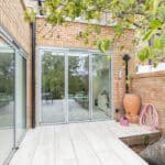 Thumbnail of http://Patio%20area%20with%20sliding%20glass%20doors,%20tree%20near%20wall,%20pink%20hose,%20large%20terracotta%20pot,%20and%20outdoor%20furniture%20visible%20inside.