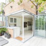 Thumbnail of http://Kitchen%20and%20dining%20room%20with%20open%20bi-fold%20glass%20doors%20leading%20to%20a%20patio.%20Brick%20exterior,%20light%20wood%20flooring,%20white%20cabinetry,%20and%20green%20plants%20visible.