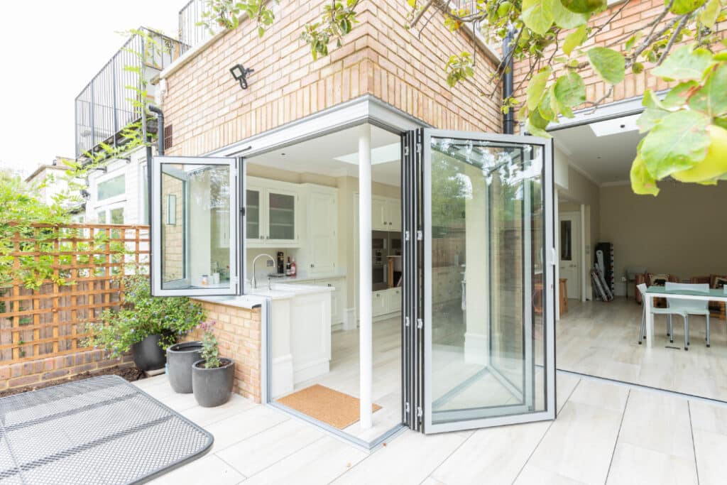 Open glass doors lead from a modern kitchen to a patio. The kitchen features white cabinetry, and the patio has potted plants and outdoor furniture.