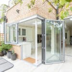 Thumbnail of http://Open%20glass%20doors%20lead%20from%20a%20modern%20kitchen%20to%20a%20patio.%20The%20kitchen%20features%20white%20cabinetry,%20and%20the%20patio%20has%20potted%20plants%20and%20outdoor%20furniture.