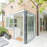 Thumbnail of http://Open%20corner%20glass%20doors%20of%20a%20brick%20house%20reveal%20a%20kitchen%20and%20dining%20area%20inside.%20The%20exterior%20patio%20features%20potted%20plants%20and%20a%20metal%20table.%20Lush%20greenery%20surrounds%20the%20outside.