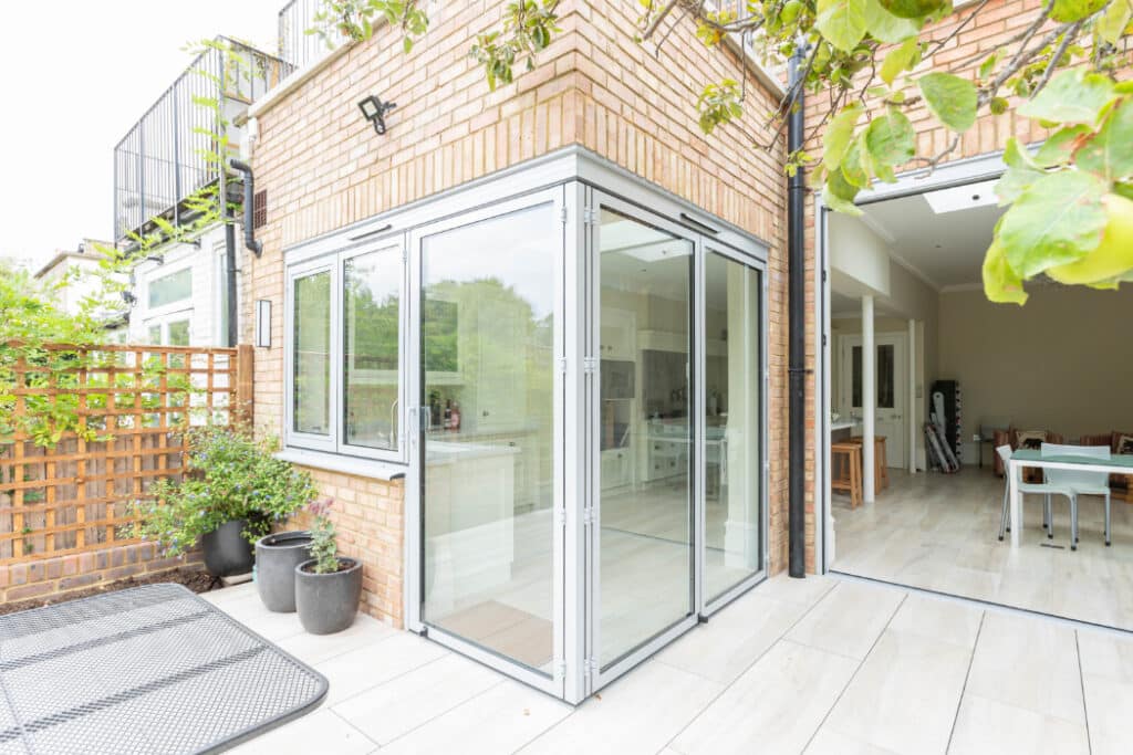Modern brick house with a glass-enclosed patio leading into an open-plan interior. Potted plants and a lattice panel are placed beside the patio.