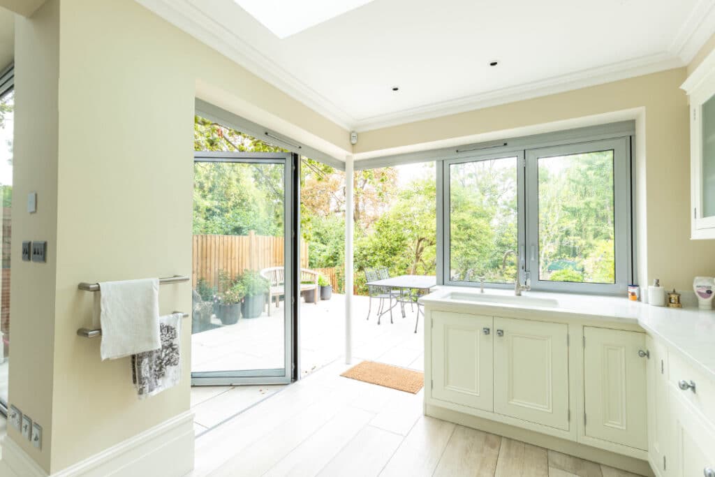 Bright kitchen with cream cabinets and light wood flooring. Glass doors open to a patio with outdoor furniture and greenery.