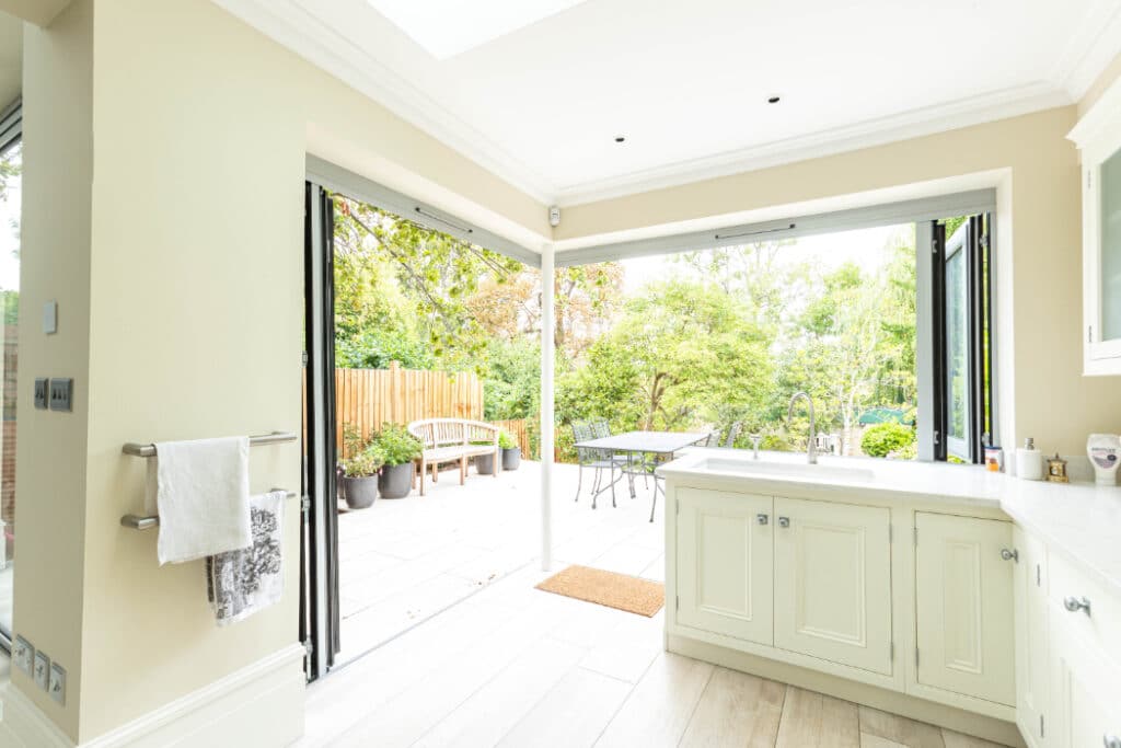 Bright kitchen with open bi-fold doors leading to a patio. White cabinets and a light wood floor are visible. The patio features outdoor furniture and is surrounded by greenery.