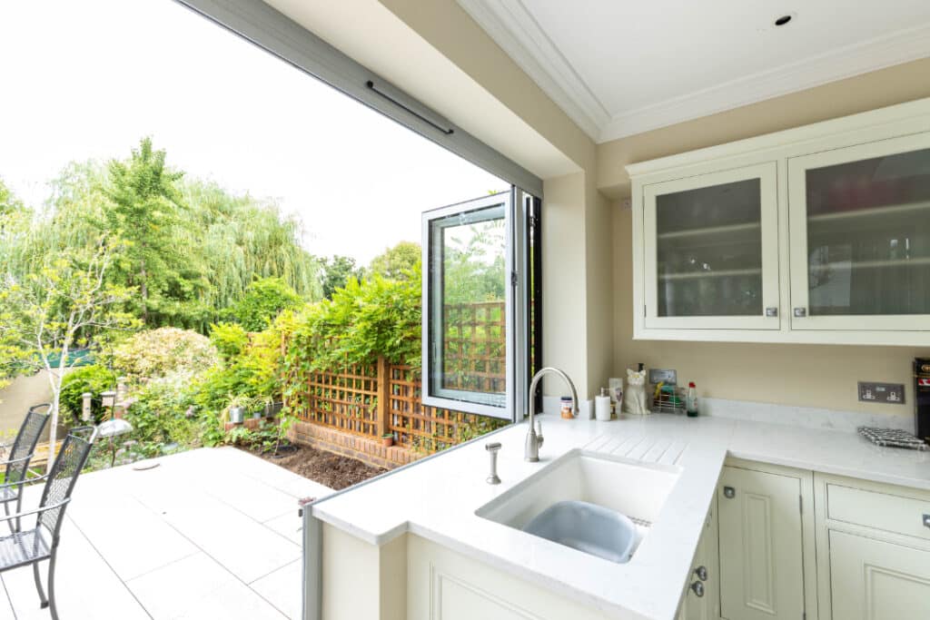 Bright kitchen with white cabinetry and countertop, open folding glass doors leading to a garden patio with outdoor furniture, and lush greenery in the background.