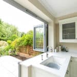 Thumbnail of http://Bright%20kitchen%20with%20white%20cabinetry%20and%20countertop,%20open%20folding%20glass%20doors%20leading%20to%20a%20garden%20patio%20with%20outdoor%20furniture,%20and%20lush%20greenery%20in%20the%20background.