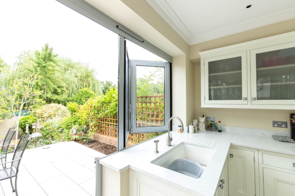 A bright kitchen with white cabinets and a sink near a large open window. The view outside shows a garden with greenery and a patio with metal chairs.