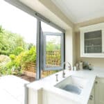 Thumbnail of http://A%20bright%20kitchen%20with%20white%20cabinets%20and%20a%20sink%20near%20a%20large%20open%20window.%20The%20view%20outside%20shows%20a%20garden%20with%20greenery%20and%20a%20patio%20with%20metal%20chairs.