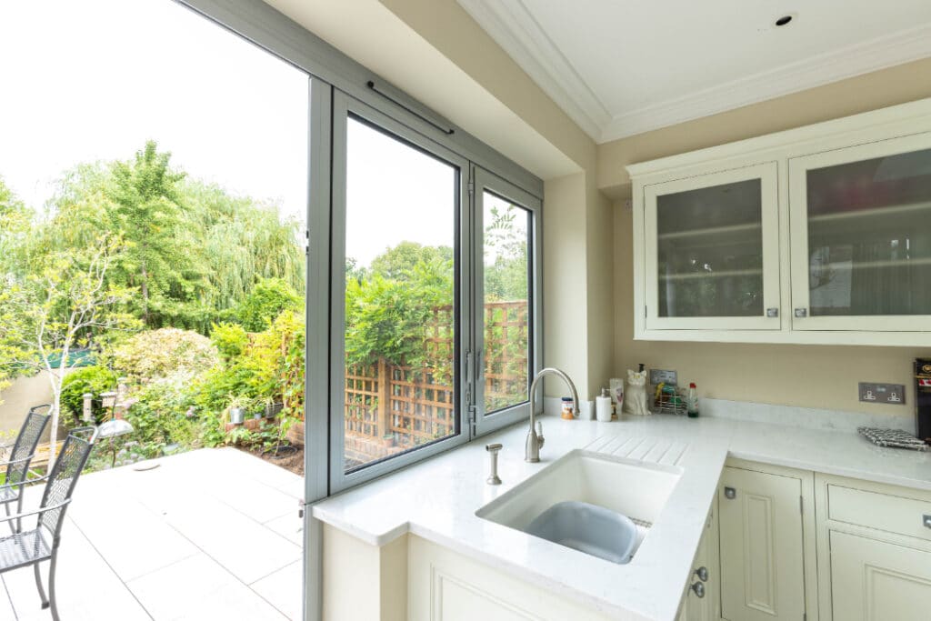 A bright kitchen with cream cabinets and a sink; large glass doors open to a garden patio with table and chairs, surrounded by greenery.