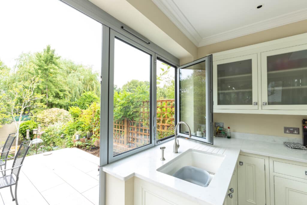 A modern kitchen with white cabinets and a marble countertop, featuring an open window overlooking a patio and a lush garden.