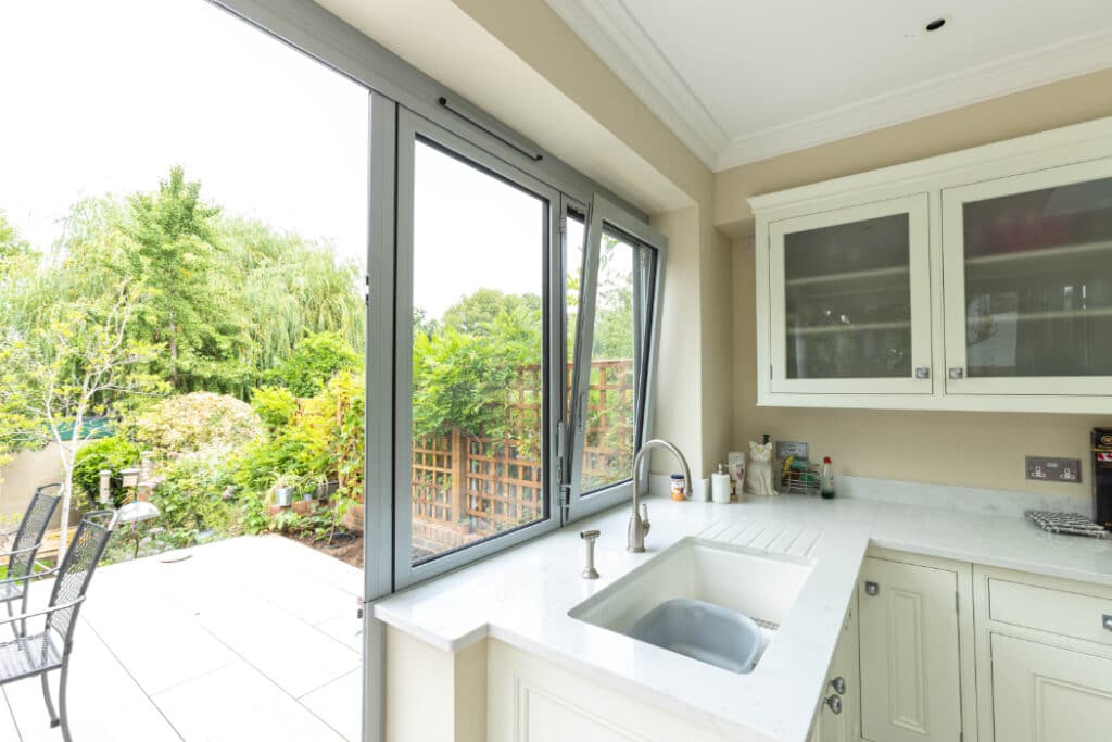 A kitchen with light-colored cabinets, a sink, and a countertop opens to a garden through a large glass sliding door.