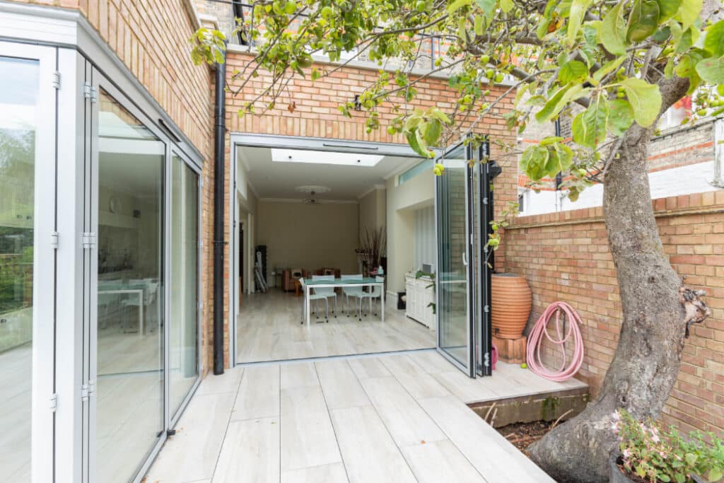 Glass doors open to a modern room with a dining table and chairs. A tree and a coiled hose are visible on the patio, alongside brick walls.