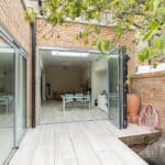 Thumbnail of http://Glass%20doors%20open%20to%20a%20modern%20room%20with%20a%20dining%20table%20and%20chairs.%20A%20tree%20and%20a%20coiled%20hose%20are%20visible%20on%20the%20patio,%20alongside%20brick%20walls.