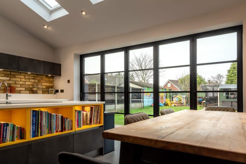 Modern kitchen with skylights, a wooden table, shelves with books, and floor-to-ceiling windows overlooking a backyard with a playhouse and trees.