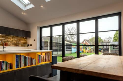 Modern kitchen with skylights, a wooden table, shelves with books, and floor-to-ceiling windows overlooking a backyard with a playhouse and trees.