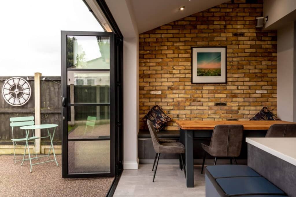 Kitchen nook with a wooden table, chairs, and a brick accent wall. Open glass door leads to an outdoor area with a green bistro set. Framed picture on the wall.