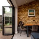 Thumbnail of http://Kitchen%20nook%20with%20a%20wooden%20table,%20chairs,%20and%20a%20brick%20accent%20wall.%20Open%20glass%20door%20leads%20to%20an%20outdoor%20area%20with%20a%20green%20bistro%20set.%20Framed%20picture%20on%20the%20wall.