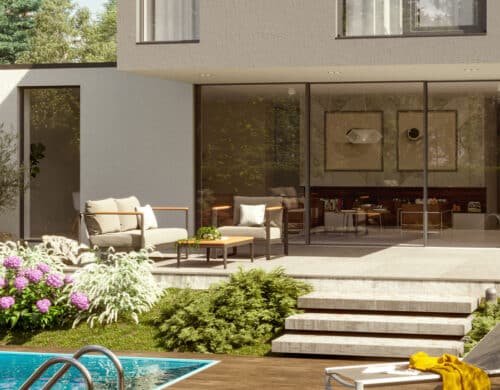 Modern house with large glass doors, patio chairs, and steps leading to a pool. Surrounded by greenery and flowers in a landscaped garden.